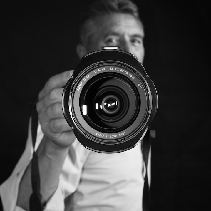 A middle aged man holding a camera out in front of him with a close up view of the lens